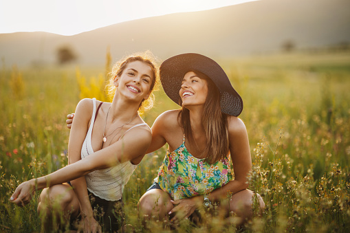 Two young cheerful female friends enjoying time together in nature, sitting in flower meadow and laughing