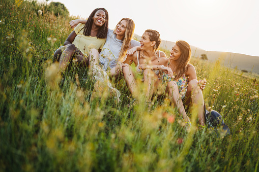 Cheerful girlfriends having fun in nature , sitting in flower meadow, talking and laughing, enjoying time together on beautiful summer day at sunset