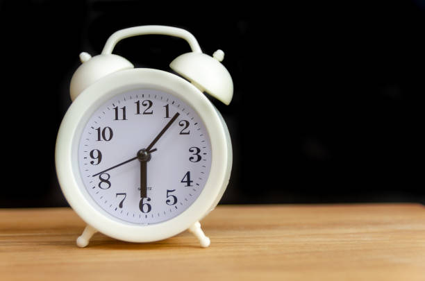 istock white alarm clock stands on a wooden table against a black wall, copy space 1262883487