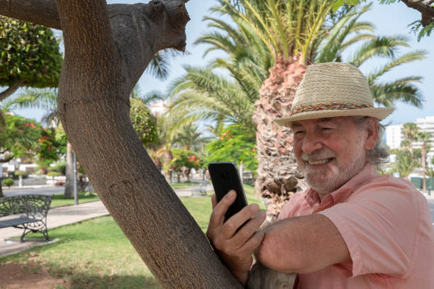 istock Cheerful bearded senior man with straw hat using mobile phone in a pleasant public park with plants and palm trees - concept of elderly active people tech and social in summer vacation 1262883817