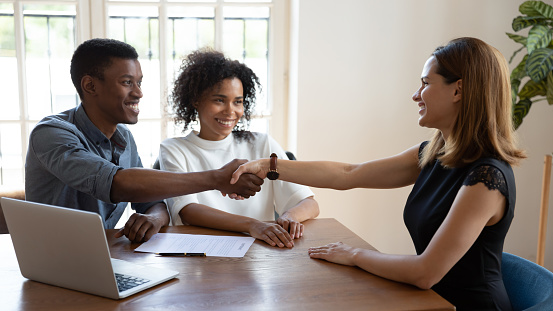 Happy smiling African married couple shake hands to mortgage insurance woman broker. Formal meeting with financial advisor, real estate agent, consulting, make deal, handshake gesture of trust concept