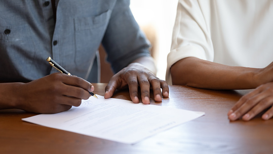 Horizontal image man holding pen put signature on agreement African couple filling form bank application taking loan, affirming rental contract, real estate purchasing, hands and table close up view