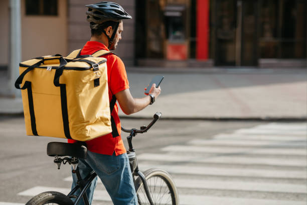 istock Delivery man on bicycle. Guy with backpack and helmet stopped at intersection and looked at phone 1262891041