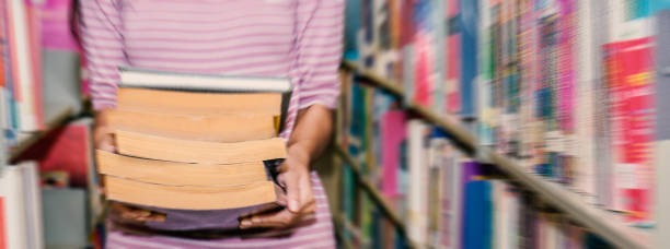 primo tempo giovane studente portando una pesante pila di libri in biblioteca. sfondo e banner del concetto di istruzione (sfocatura) - student effort book carrying foto e immagini stock