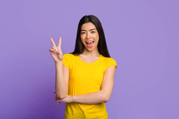 istock Cheerful Asian Girl Gesturing Peace While Posing Over Purple Studio Background 1262894999