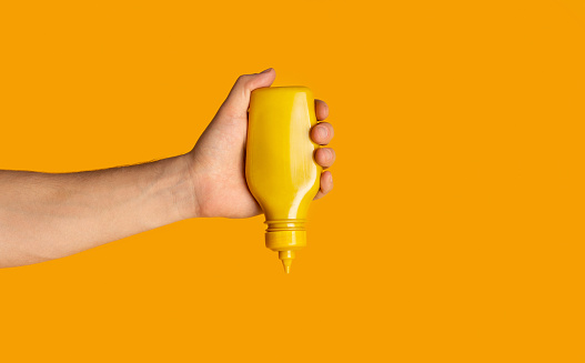 Male hand squeezing bottle with mustard on orange background, close up. Space for text