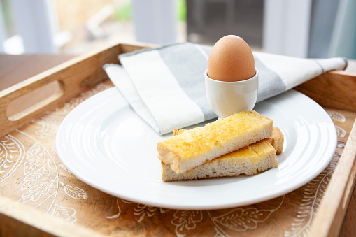 Unbroken egg in an egg cup with buttered toast soldiers on a white plate with a napkin on a wooden tray