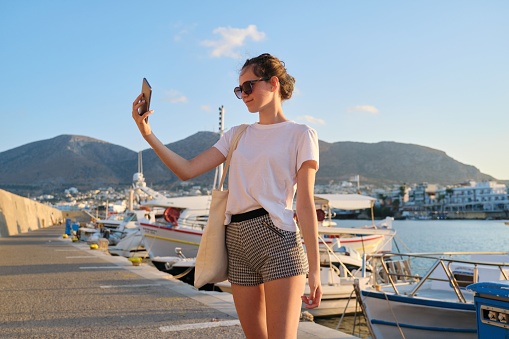 Fashionable beautiful girl teenager walking on pier, sunset on sea, moored yachts in bay, mountain landscape background. Girl in sunglasses, shorts takes selfie on smartphone.