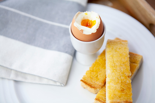 A broken boiled egg in an egg cup with buttered toast soldiers on a white plate with a napkin on a wooden tray viewed from a high angle