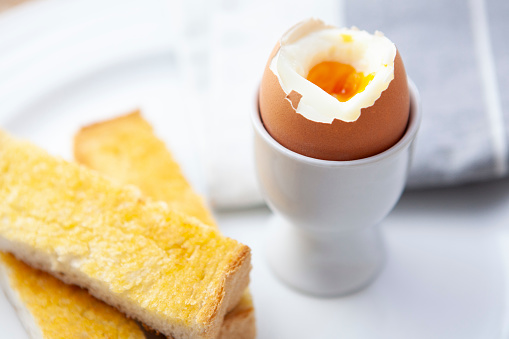 A broken boiled egg in an egg cup with buttered toast soldiers on a white plate with a napkin