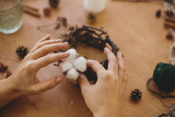 istock Florist making rustic christmas candle holder. Hands holding cotton and making rustic festive decoration on wooden table with branches and pine cones. Holiday workshop 1262901559
