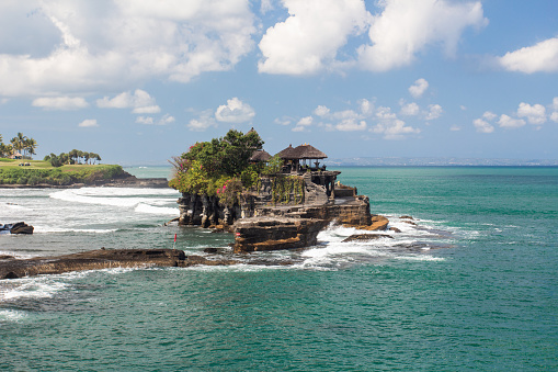 Aerial view of Tanah Lot temple on cliff in Indian ocean. Waves are breaking on a rock cliff off the coast of Bali. Hindu temple built on a rock in the sea. Lush tropical vegetation on top of a cliff.