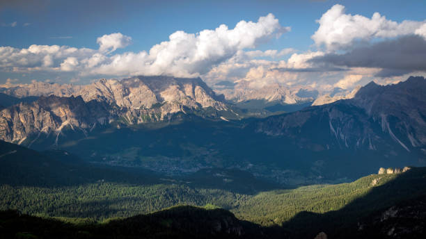 Wide angle view of forested valley in Italian Dolomites. Distant peaks are lit by warm sunlight; town in the valley remains in shadow. stock photo