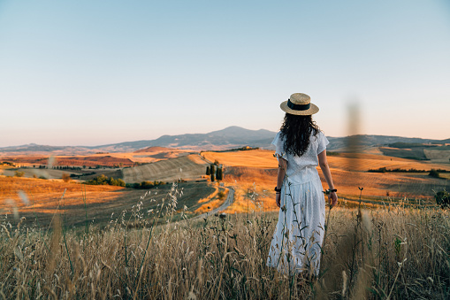 Young woman admiring sunset in a wheat field in Tuscany. She's wearing white clothes and a straw hat.
