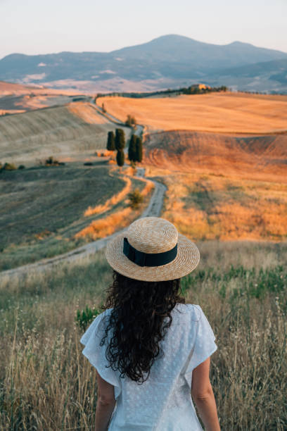 Young woman admiring sunset in a wheat field in Tuscany Young woman admiring sunset in a wheat field in Tuscany. She's wearing white clothes and a straw hat. crete senesi photos stock pictures, royalty-free photos & images