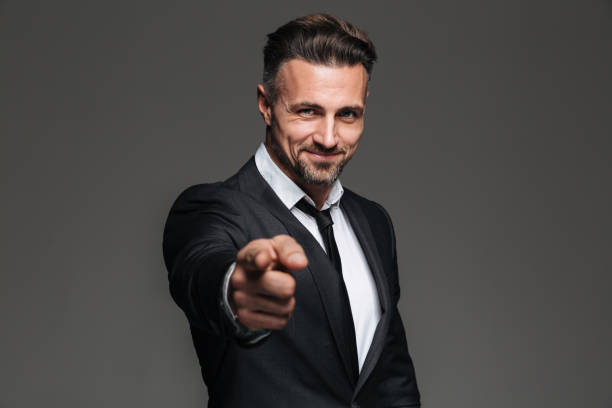 Portrait of a confident businessman pointing finger at camera Photo of attractive businessman in suit smiling and pointing index finger on camera isolated over dark gray background index finger stock pictures, royalty-free photos & images