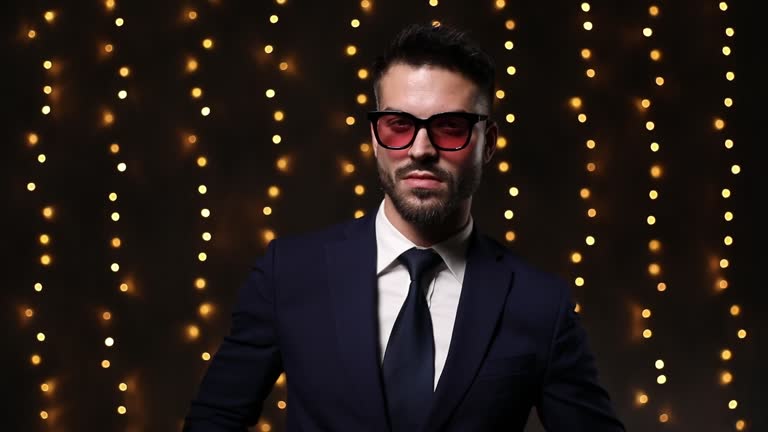 sexy young man in suit wearing sunglasses, pulling down and fixing suit, crossing arms and smiling, holding hand to face and touching chin and beard, adjusting suit, moving in a side view position and walking away on lights background