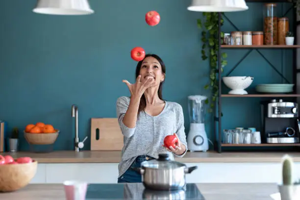Shot of funny young woman juggling with three red apples in the kitchen at home.