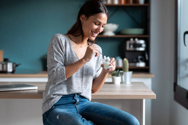 自宅の台所でスツールに座りながらヨーグルトを食べる笑顔の若い女性。 - ヨーグルト ストックフォトと画像