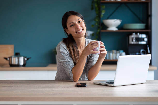 giovane donna sorridente che guarda la macchina fotografica mentre tiene una tazza di caffè e lavora con il laptop in cucina a casa. - smart casual foto e immagini stock