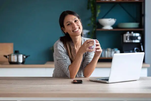 Shot of smiling young woman looking at camera while holding a cup of coffee and working with laptop in the kitchen at home.