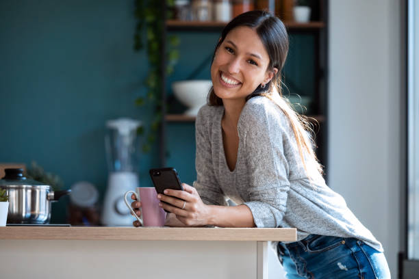 istock Smiling young woman looking at camera while using her mobile phone and drinking a cup of coffee in the kitchen at home. 1262919109