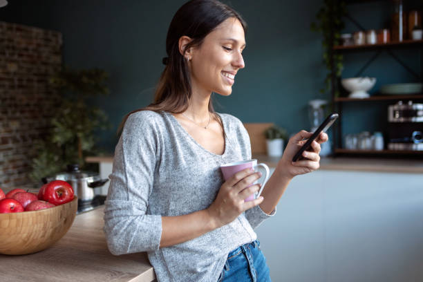 istock Smiling young woman using her mobile phone while drinking a cup of coffee in the kitchen at home. 1262919163
