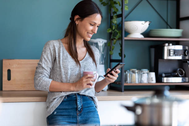 istock Smiling young woman using her mobile phone while drinking a cup of coffee in the kitchen at home. 1262919214