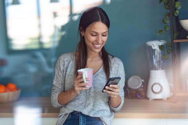 istock Smiling young woman using her mobile phone while drinking a cup of coffee in the kitchen at home. 1262919343