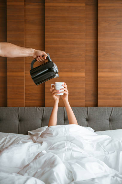 adolescente debaixo de cobertor em cama branca no quarto acordou e chá está sendo derramado para ela. café da manhã na cama, manhã preguiçosa - five oclock tea - fotografias e filmes do acervo