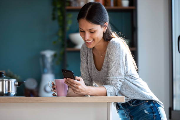 istock Smiling young woman using her mobile phone while drinking a cup of coffee in the kitchen at home. 1262919479