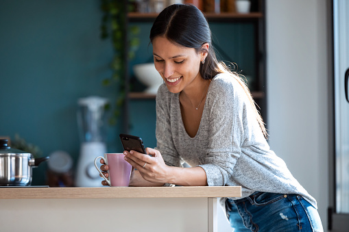 Shot of smiling young woman using her mobile phone while drinking a cup of coffee in the kitchen at home.