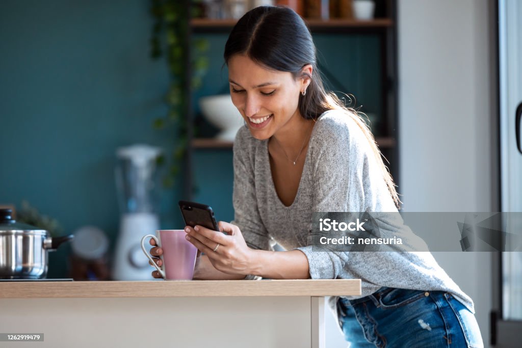 Smiling young woman using her mobile phone while drinking a cup of coffee in the kitchen at home. Shot of smiling young woman using her mobile phone while drinking a cup of coffee in the kitchen at home. Mobile Phone Stock Photo