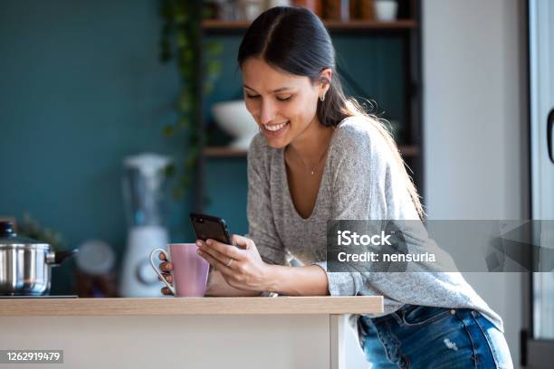Smiling Young Woman Using Her Mobile Phone While Drinking A Cup Of Coffee In The Kitchen At Home Stock Photo - Download Image Now