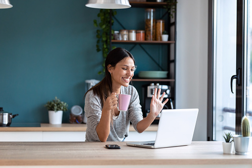 Shot of smiling young woman waving through the laptop web camera while holding a cup of coffee in the kitchen at home.