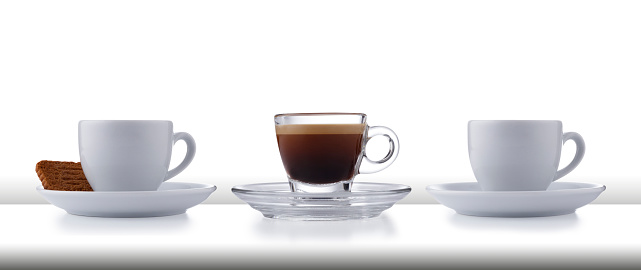 A row of 3 glass and white expresso cups and saucers full of smooth expresso coffee, on a white style bar or table top with a white background