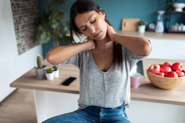 Tired young woman suffering neck pain while sitting on the stool in the kitchen at home. Shot of tired young woman suffering neck pain while sitting on the stool in the kitchen at home. muscle and joint aches stock pictures, royalty-free photos & images