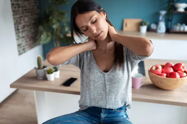 Photo of Tired young woman suffering neck pain while sitting on the stool in the kitchen at home.