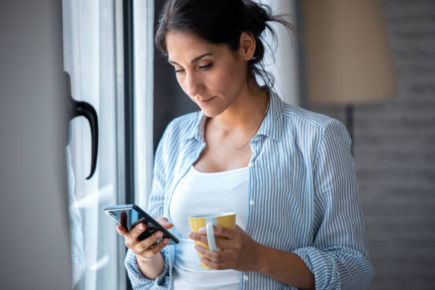 istock Pretty young woman using her mobile phone while drinking coffee near to the window in the living room at home. 1262946751