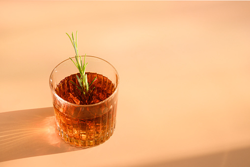One Glass of cold whiskey garnish rosemary on beige background with sunny shadow. Copy space.
