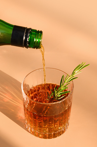 Pouring whiskey in glass with ice cube on beige background with sunny shadow. Close up. Vertical format.