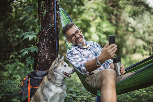 Mature man at forest. Sitting at hammock and enjoying in nature with his dog.