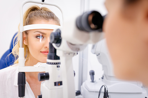 Pretty young woman leaning chin on optical instrument and having her eyes examed by the doctor