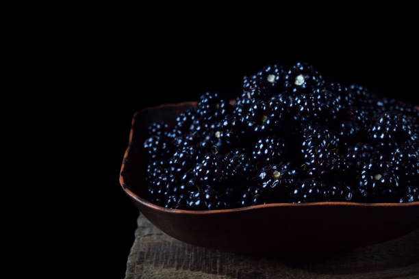 Blackberries in a clay bowl on the edge of the wooden table against the black background
