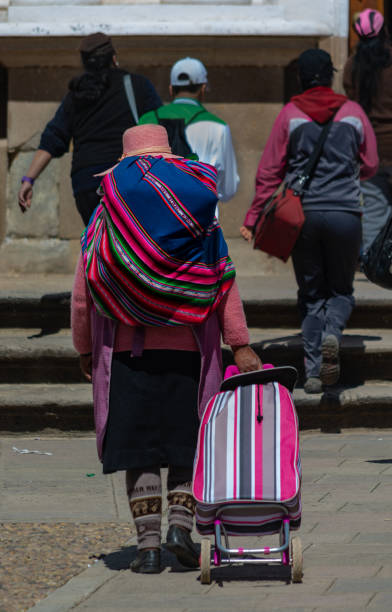 una mujer boliviana lleva un "aguayo" y un carrito de bolsa de la compra en copacabana, bolivia - bolivia copacabana bolivian ethnicity lake titicaca fotografías e imágenes de stock