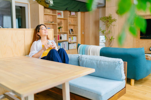 joven mujer sentada en la mesa en la sala de estar sosteniendo bebida caliente y disfrutando de su tiempo - tea women beauty pampering fotografías e imágenes de stock