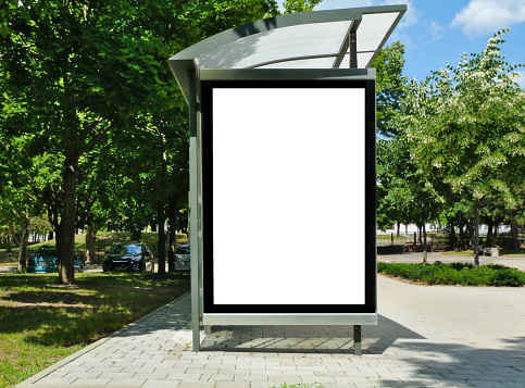composite raster image of bus shelter at a bus stop of transparent clear glass and aluminum frame structure in green street setting with trees and street in the background. milky white poster ad and banner display glass. white light box. copy space.