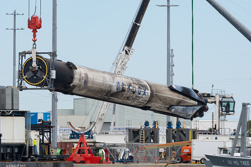 July 25, 2020 - Cape Canaveral, USA: A SpaceX Falcon 9 booster rocket is being hoisted by 2 cranes. It will be loaded to the white flatbed rocket transporter in the photo. It was brought into port on the drone ship/barge in the photo at bottom left  and will be refurbished and reused. The SpaceX ship Miss Tree is also on the photo at bottom right. The view is from the popular public Cove area on the south side of Port Canaveral.