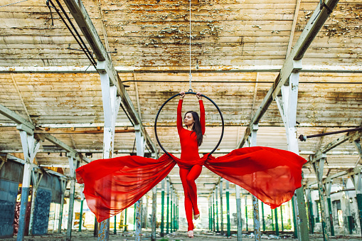 Beautiful and flexible young woman performing and dancing on aerial hoop in abandoned warehouse. She is playing makes some nice and elegant moves. Red one-piece costume reveals her perfect body shaped with sports and good habits.