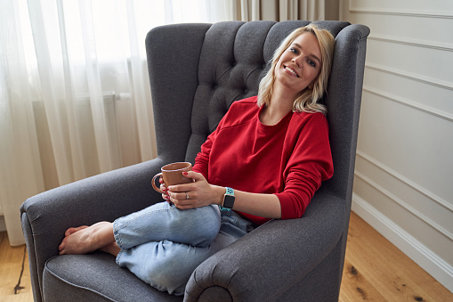 Cheerful pretty woman smiling while sitting comfortably in her favorite armchair with a mug of tea